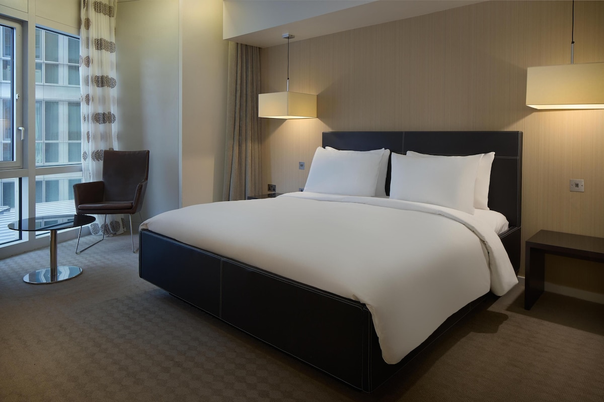 A modern hotel room with a large bed, a chair, a small table, and a window with curtains.