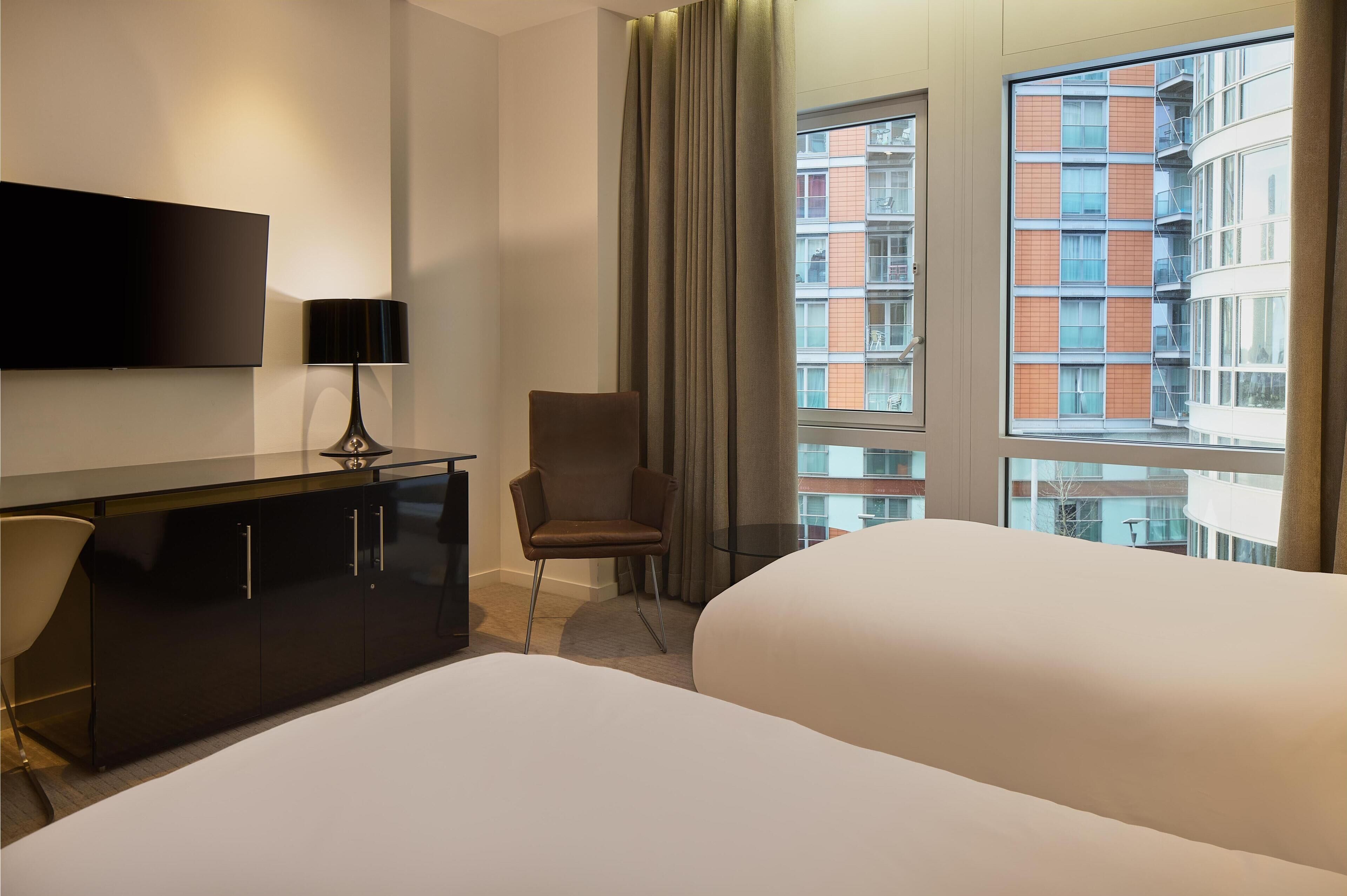 Ett hotellrum med en platt-TV, ett skrivbord med en lampa, en stol och två sängar.