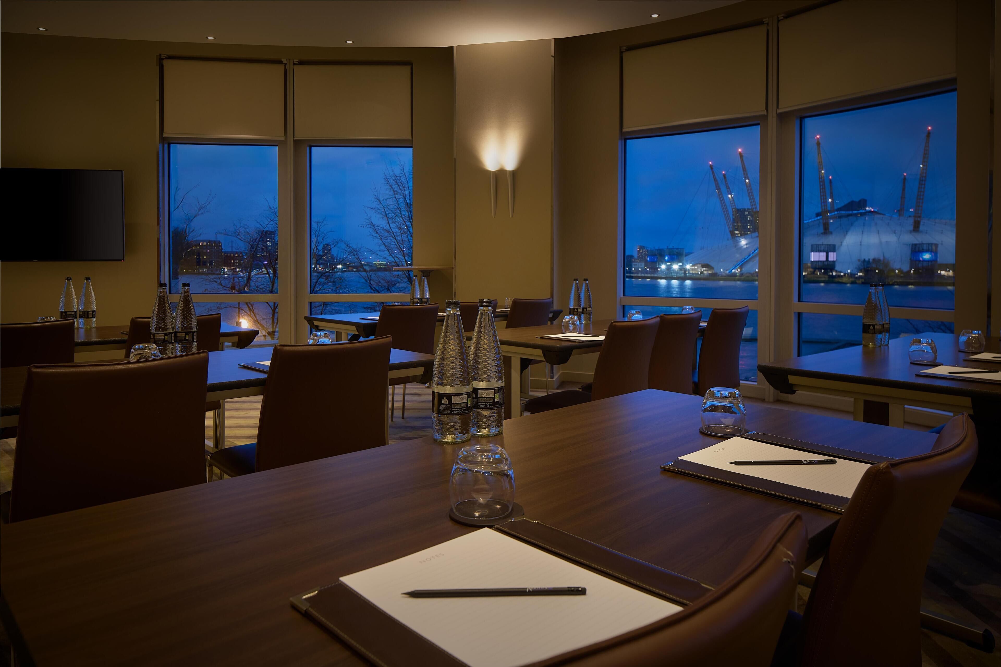 A restaurant with tables set for dining, large windows with city views, and a flat-screen TV mounted on the wall.