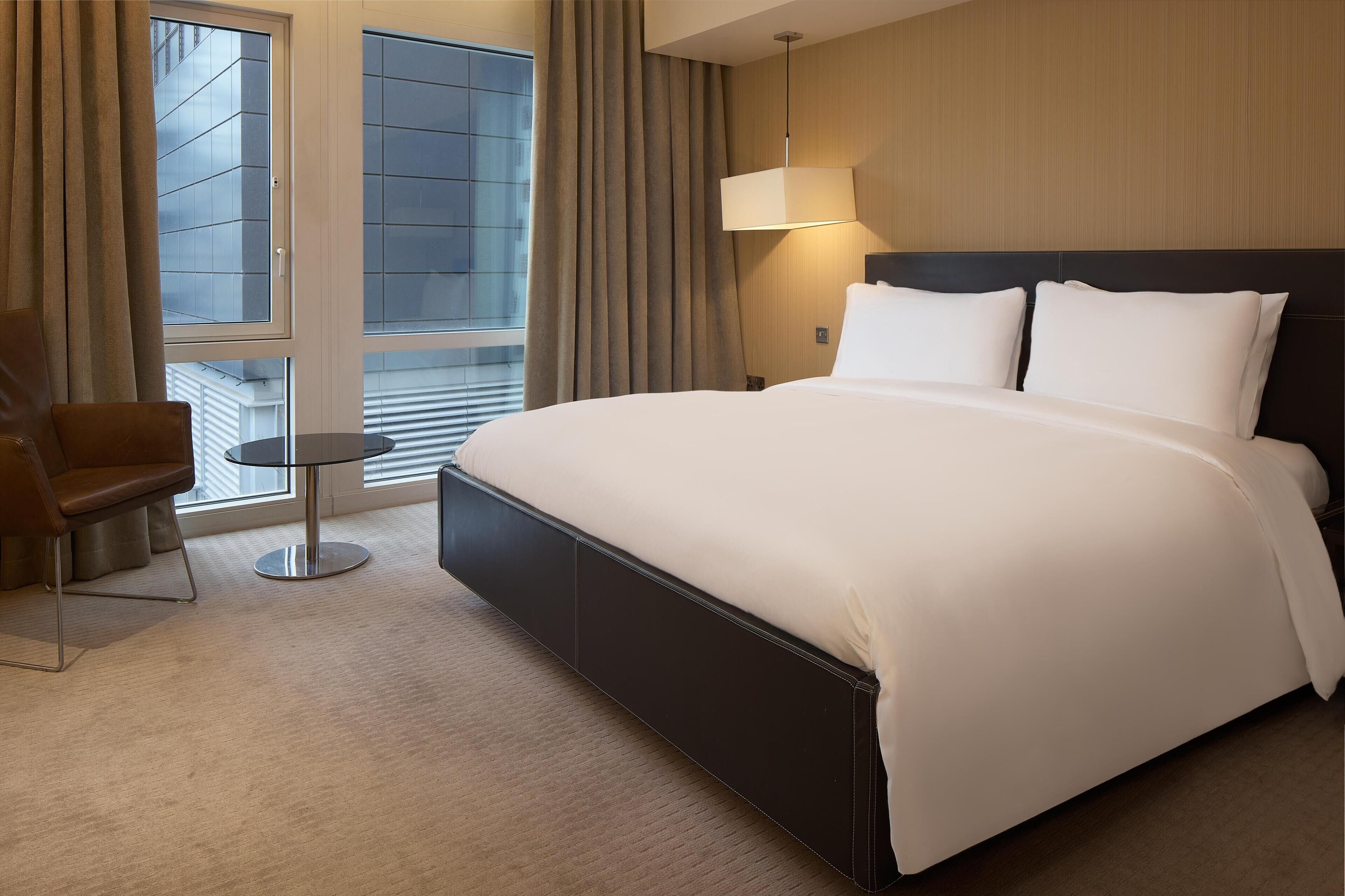 A hotel room with a large bed, a chair, a small table, and a window with curtains.