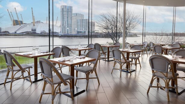 A restaurant with tables and wicker chairs, overlooking a river and construction cranes. - Radisson Blu Hotel, London Canary Wharf East (London)