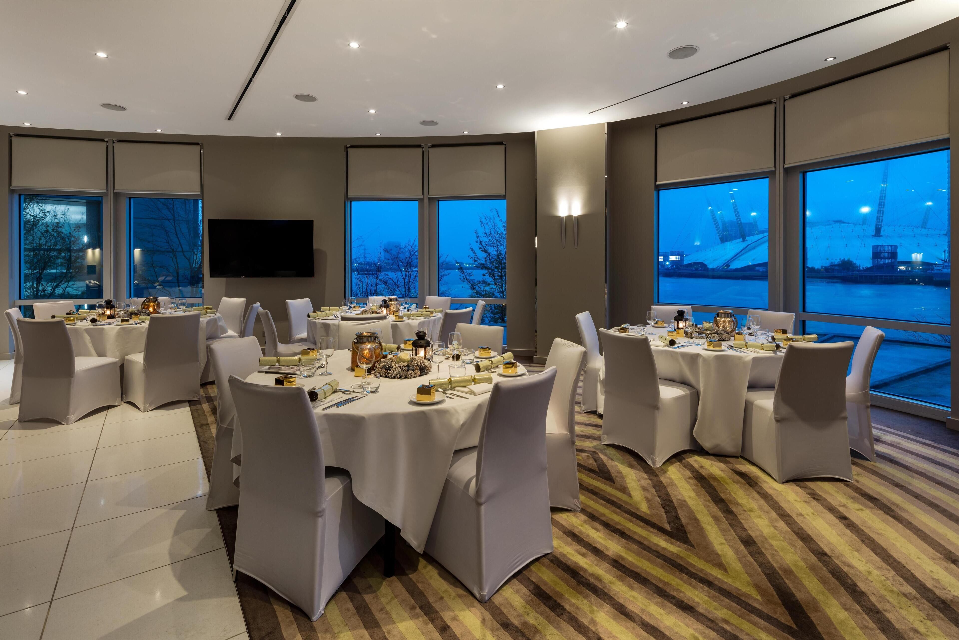 Un comedor moderno con varias mesas redondas, sillas blancas y un amplio ventanal con vista a un puente y al agua.