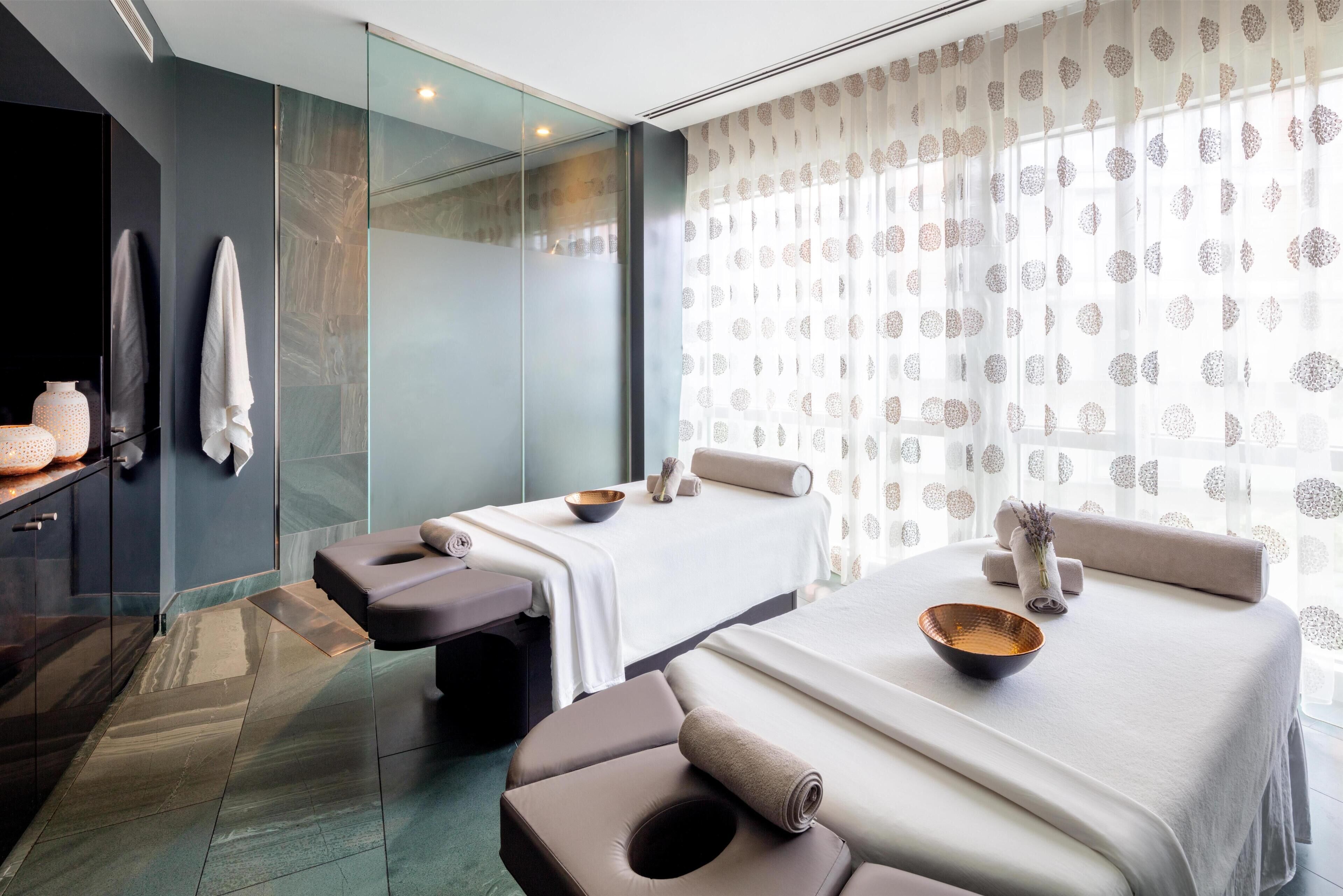 A modern spa room with two massage tables, a glass shower enclosure, and a large window with patterned curtains.