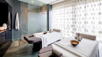 A modern spa room with two massage tables, a glass shower enclosure, and a large window with patterned curtains.