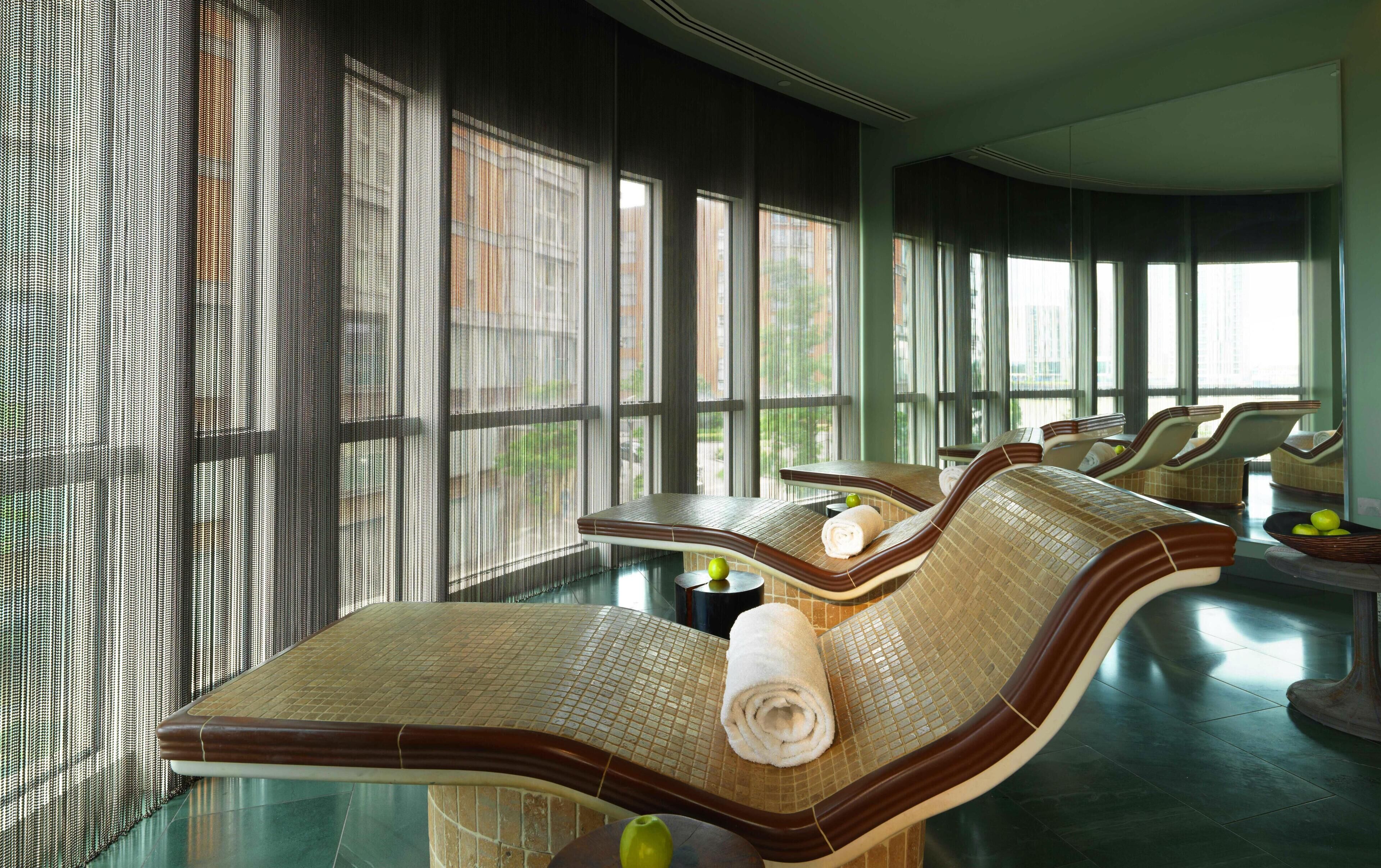 Une salle de spa moderne avec de grandes fenêtres, un espace de détente avec des fauteuils et une petite table sur laquelle sont posés une serviette et des fruits.