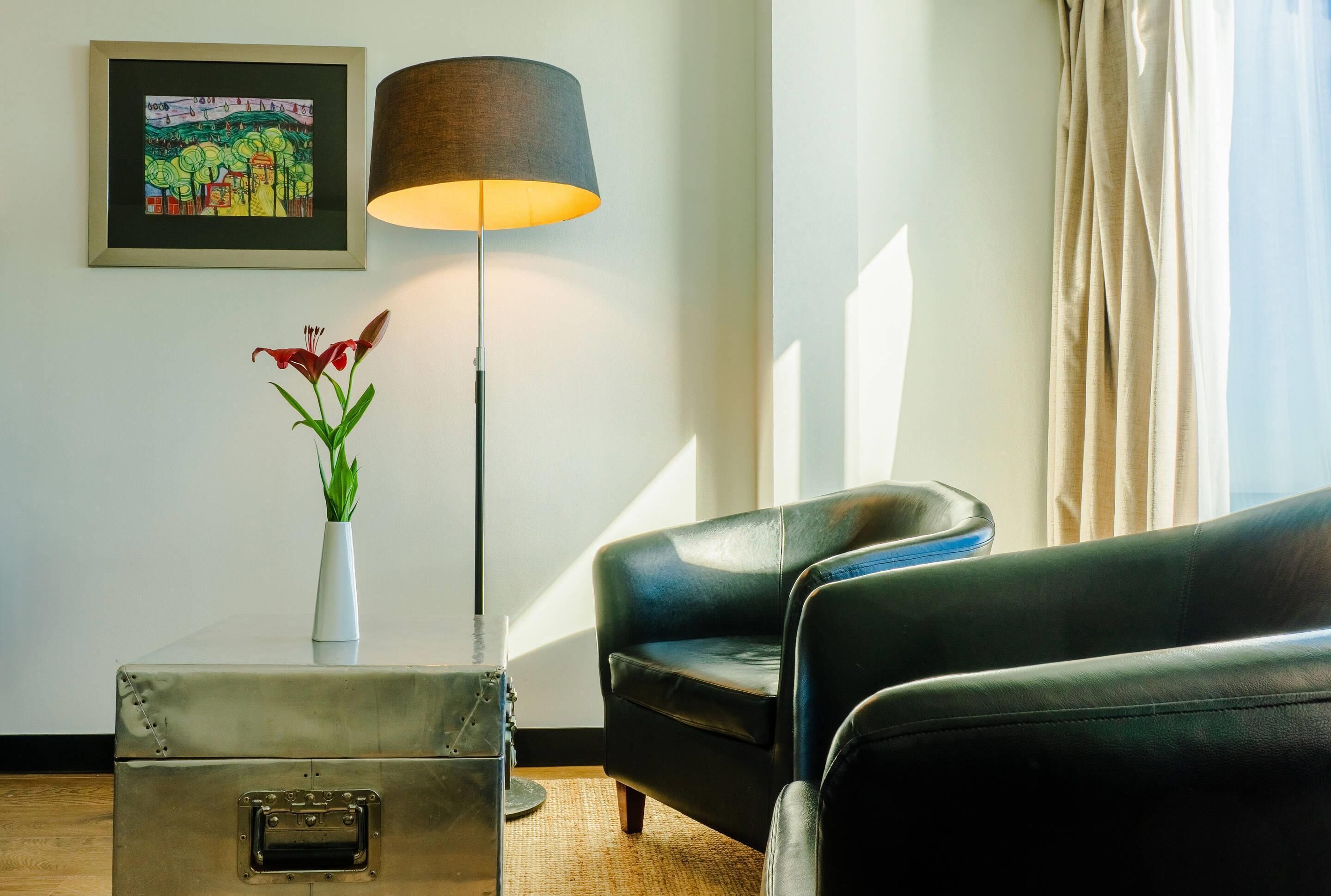Um quarto com um sofá de couro preto, uma luminária de chão, um quadro emoldurado e um vaso com flores sobre uma mesa.