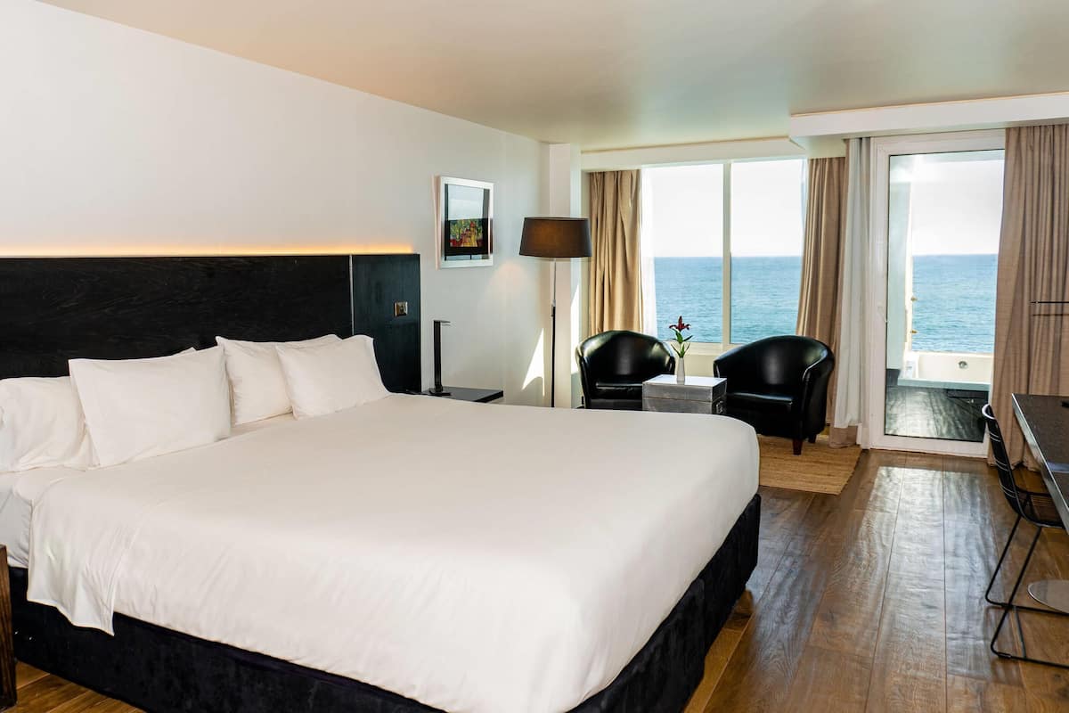 A large bed with white linens, a black headboard, a nightstand with a lamp, a chair, a small table, and a view of the sea through a large window with curtains.