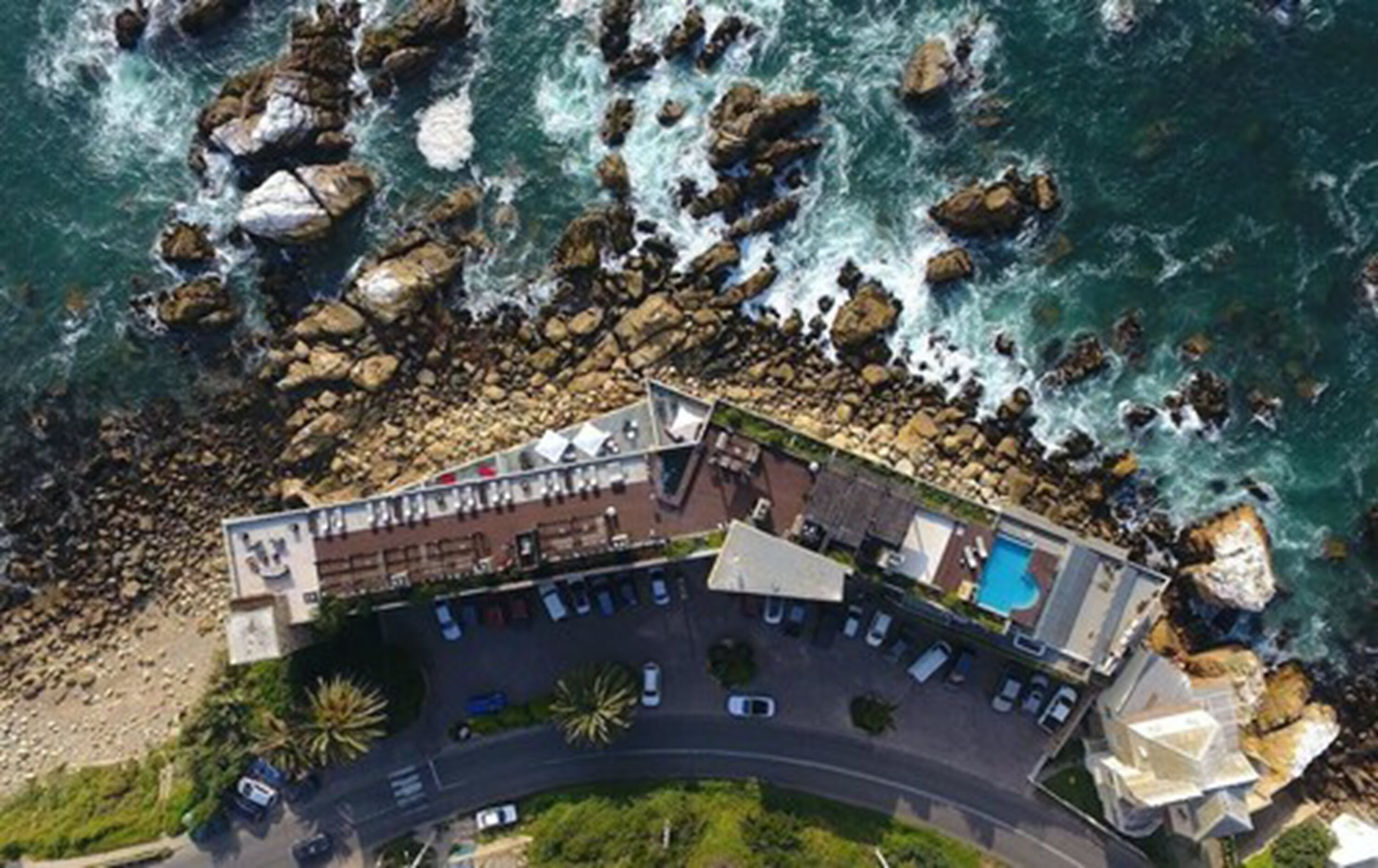 Vista aérea de un hotel costero con piscina, estacionamiento y un litoral rocoso.