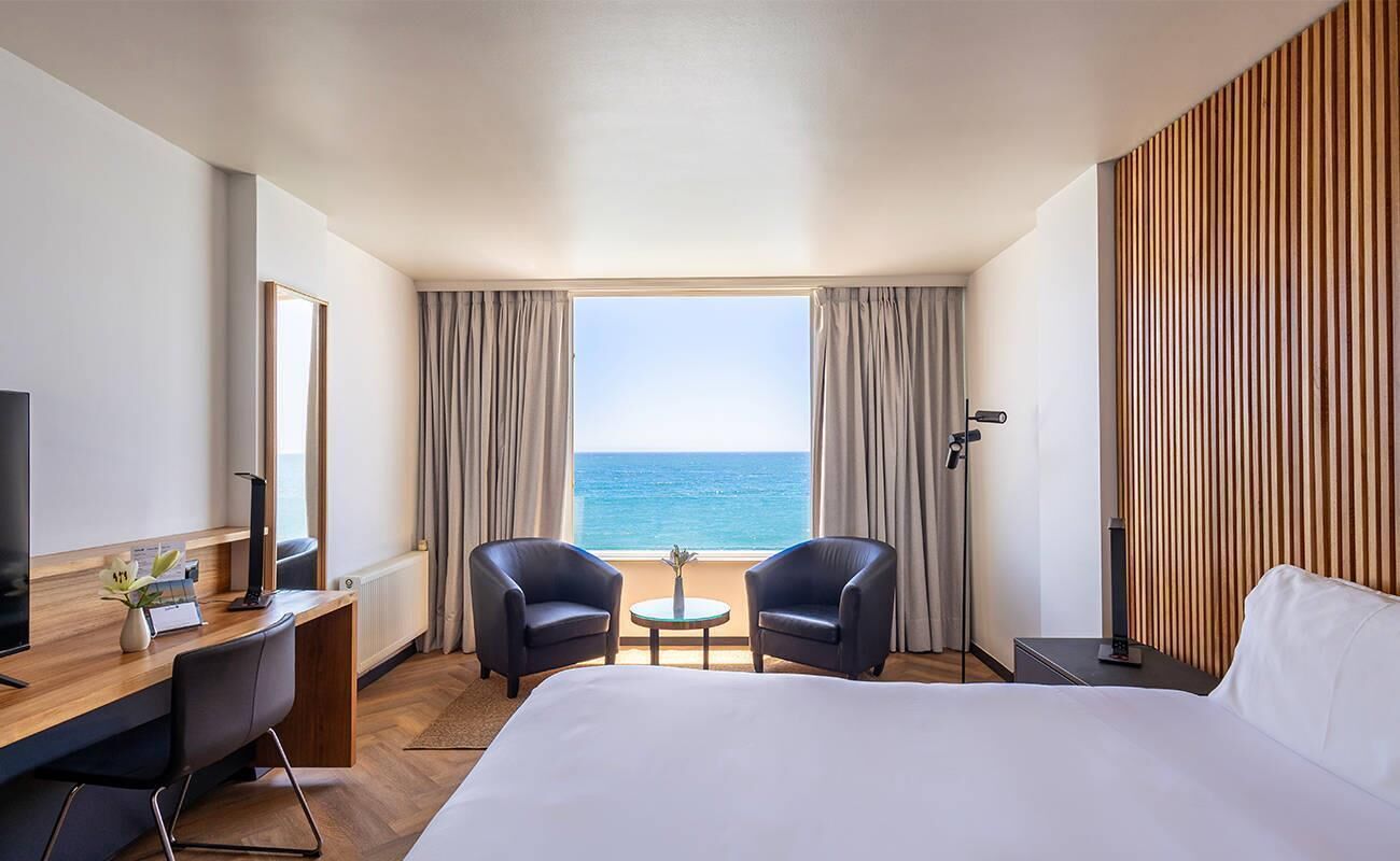 Una habitación de hotel con cama, dos sillones, una mesita, un escritorio con computadora, televisor y vista al mar.
