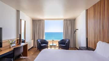 Una habitación de hotel con cama, dos sillones, una mesita, un escritorio con computadora, televisor y vista al mar.