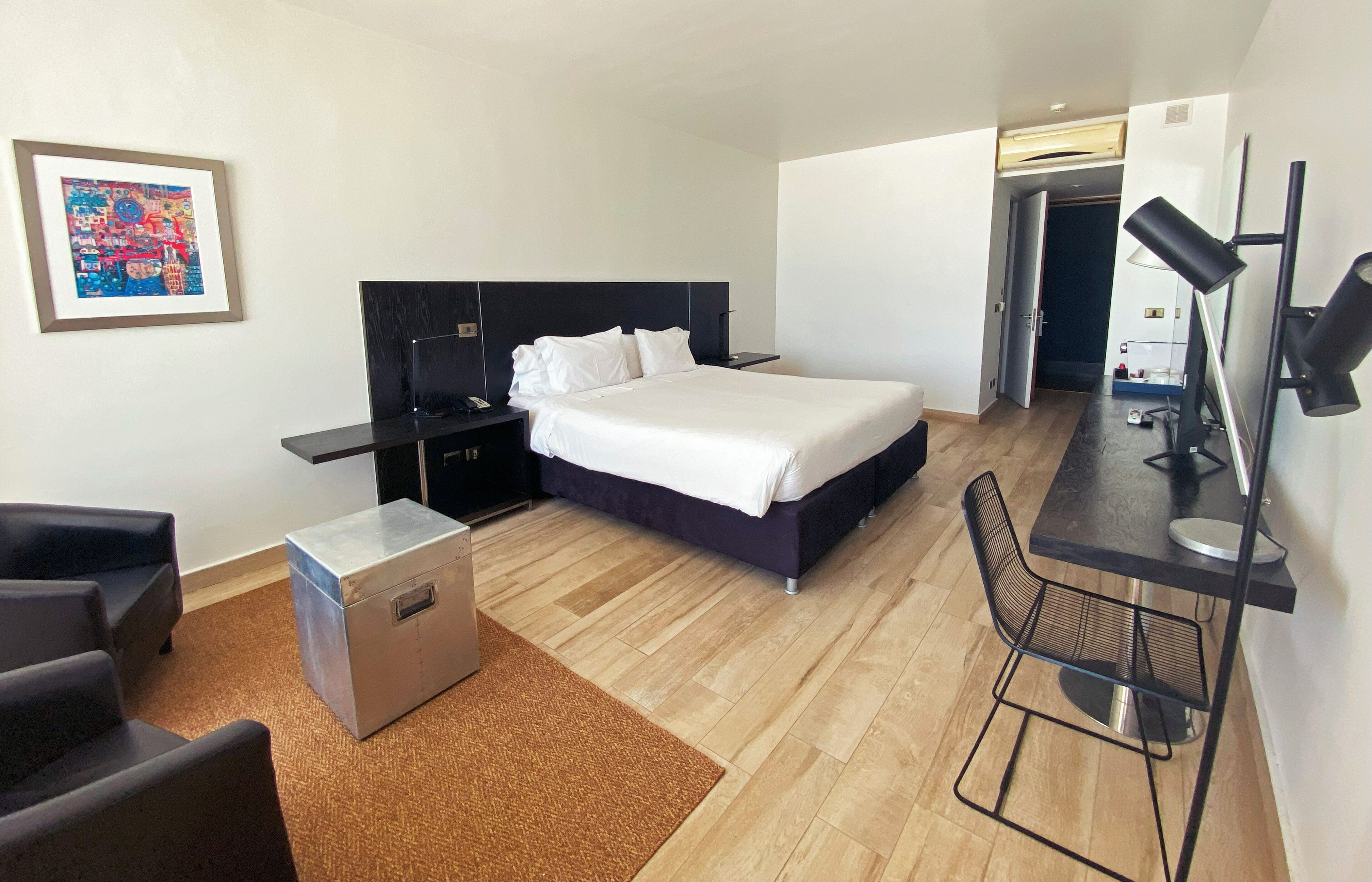 A modern hotel room with a bed, desk, chair, and a small storage unit.