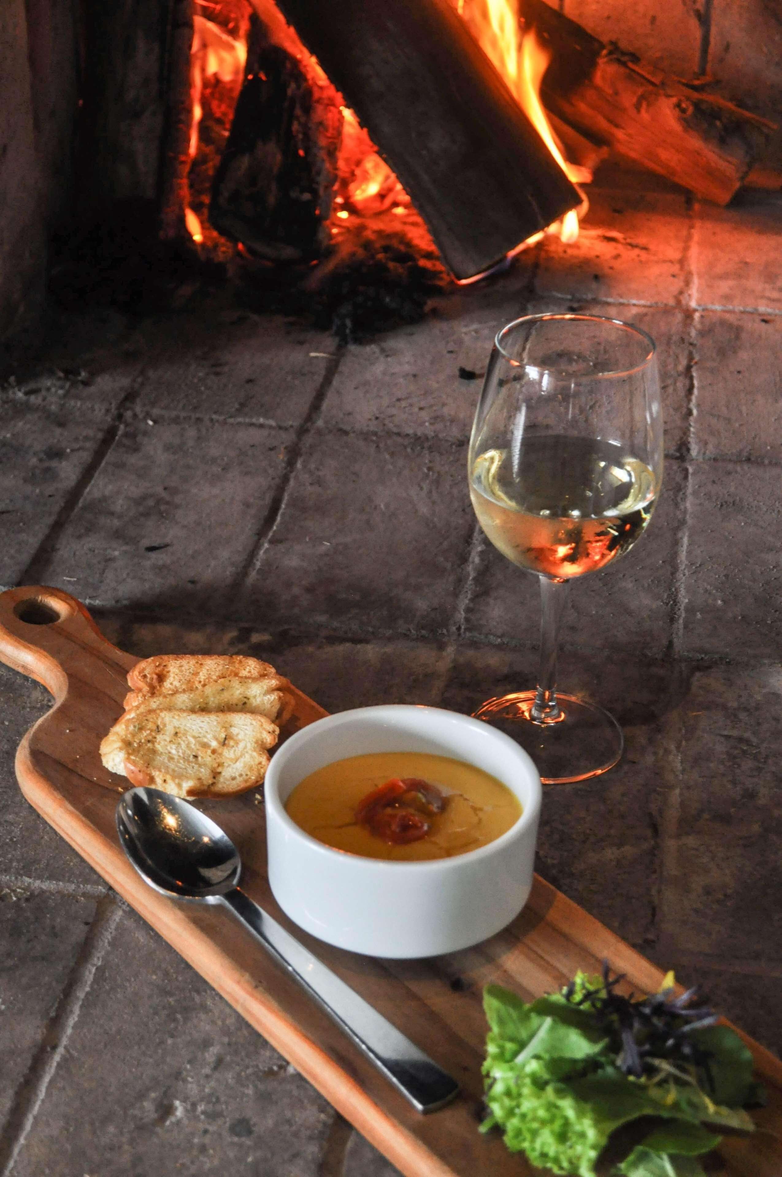 A bowl of soup with a spoon and a glass of white wine on a wooden board, with a fire in the background.