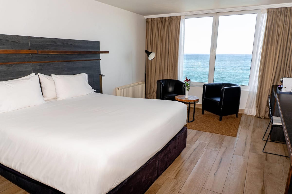 A hotel room with a large bed, a desk, a chair, and a view of the sea.