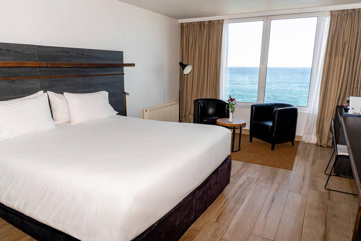 A hotel room with a large bed, a desk, a chair, and a view of the sea.