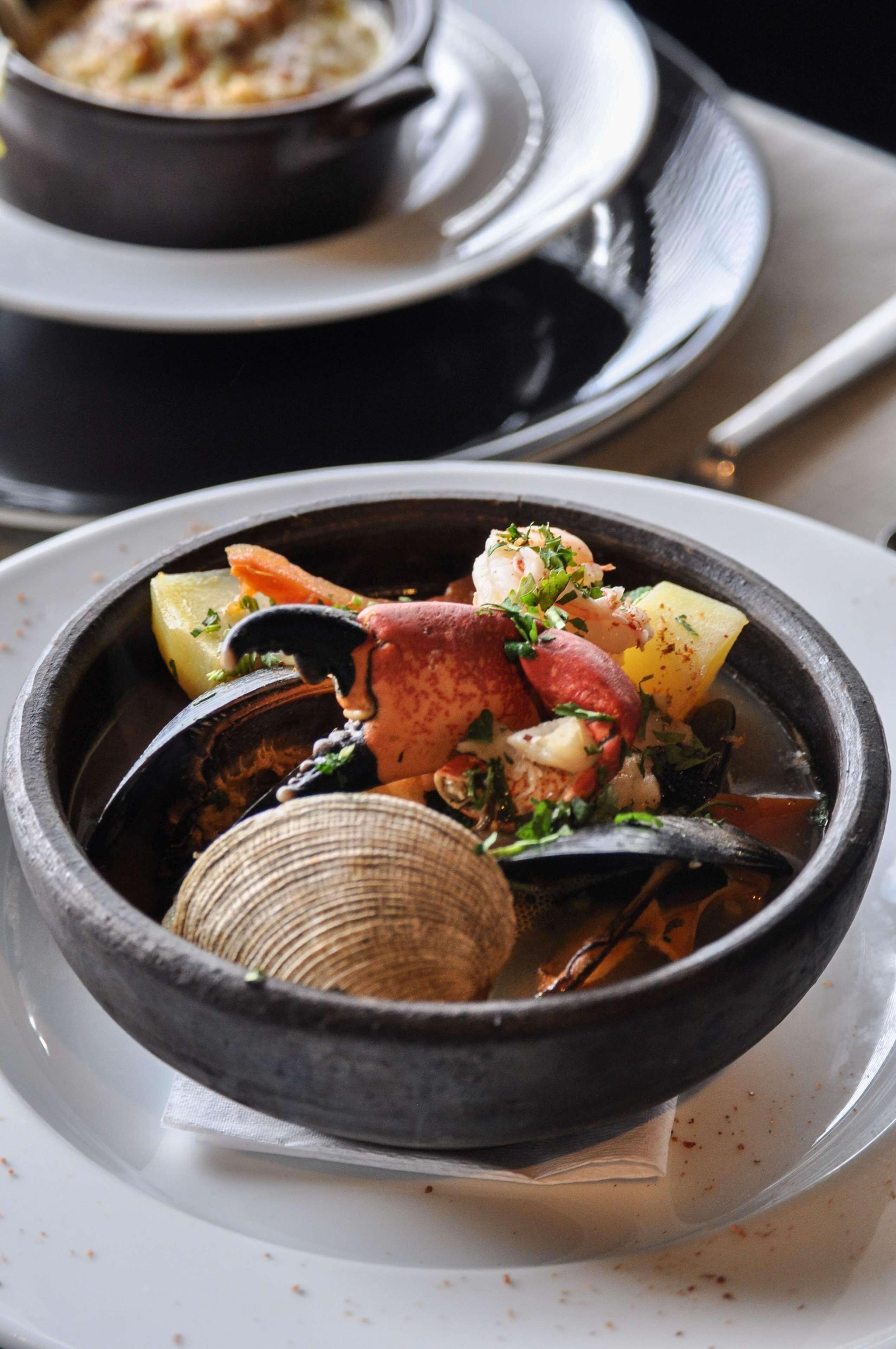 A bowl of seafood stew with clams, mussels, and shrimp, served in a black bowl on a white plate.