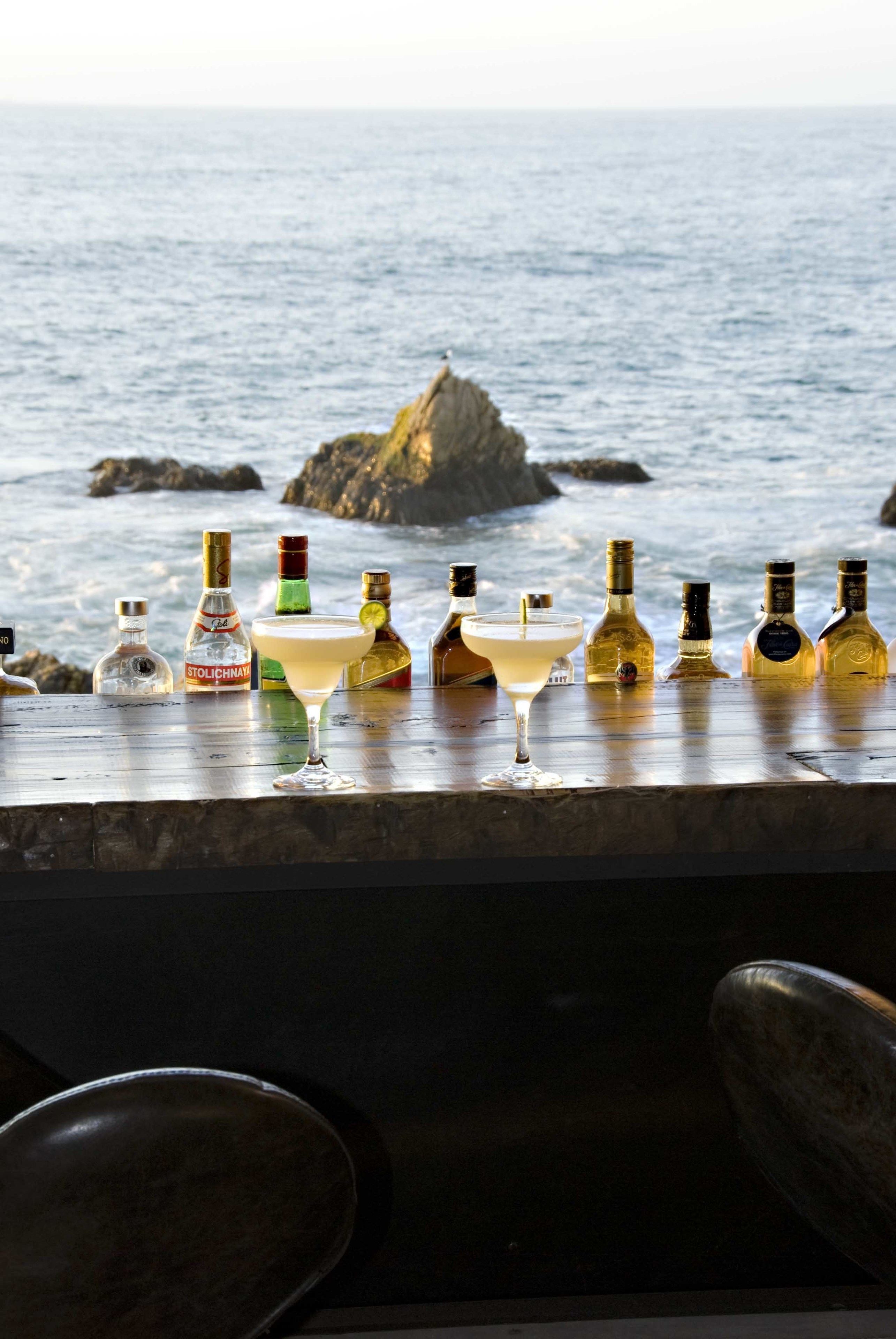 Un bar con diversas botellas y una copa de martini, con vista al océano y formaciones rocosas al fondo.