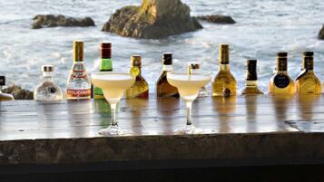 Un bar con diversas botellas y una copa de martini, con vista al océano y formaciones rocosas al fondo.