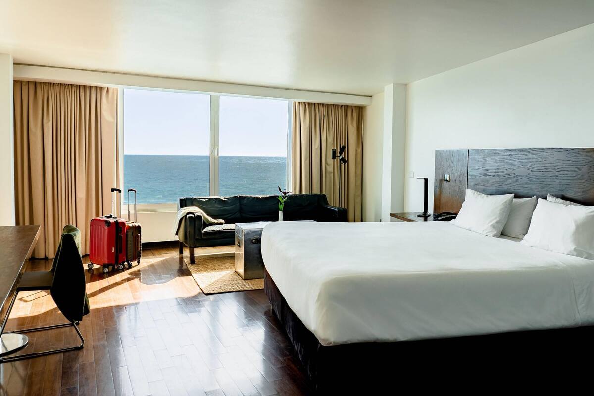 A hotel room with a large bed, a desk, a chair, a sofa, and a view of the sea.