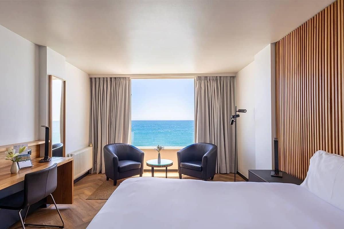 A hotel room with a bed, two armchairs, a small table, a desk with a computer, a TV, and a view of the sea.