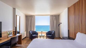 A hotel room with a bed, two armchairs, a small table, a desk with a computer, a TV, and a view of the sea.