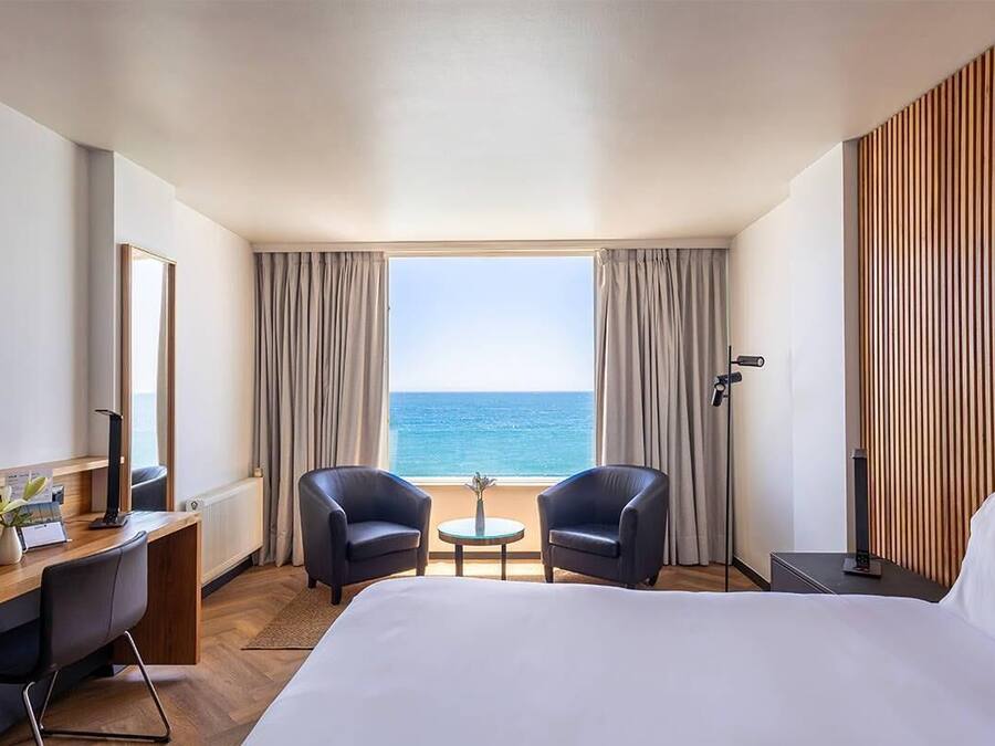 A hotel room with a bed, two armchairs, a small table, a desk with a computer, a TV, and a view of the sea.