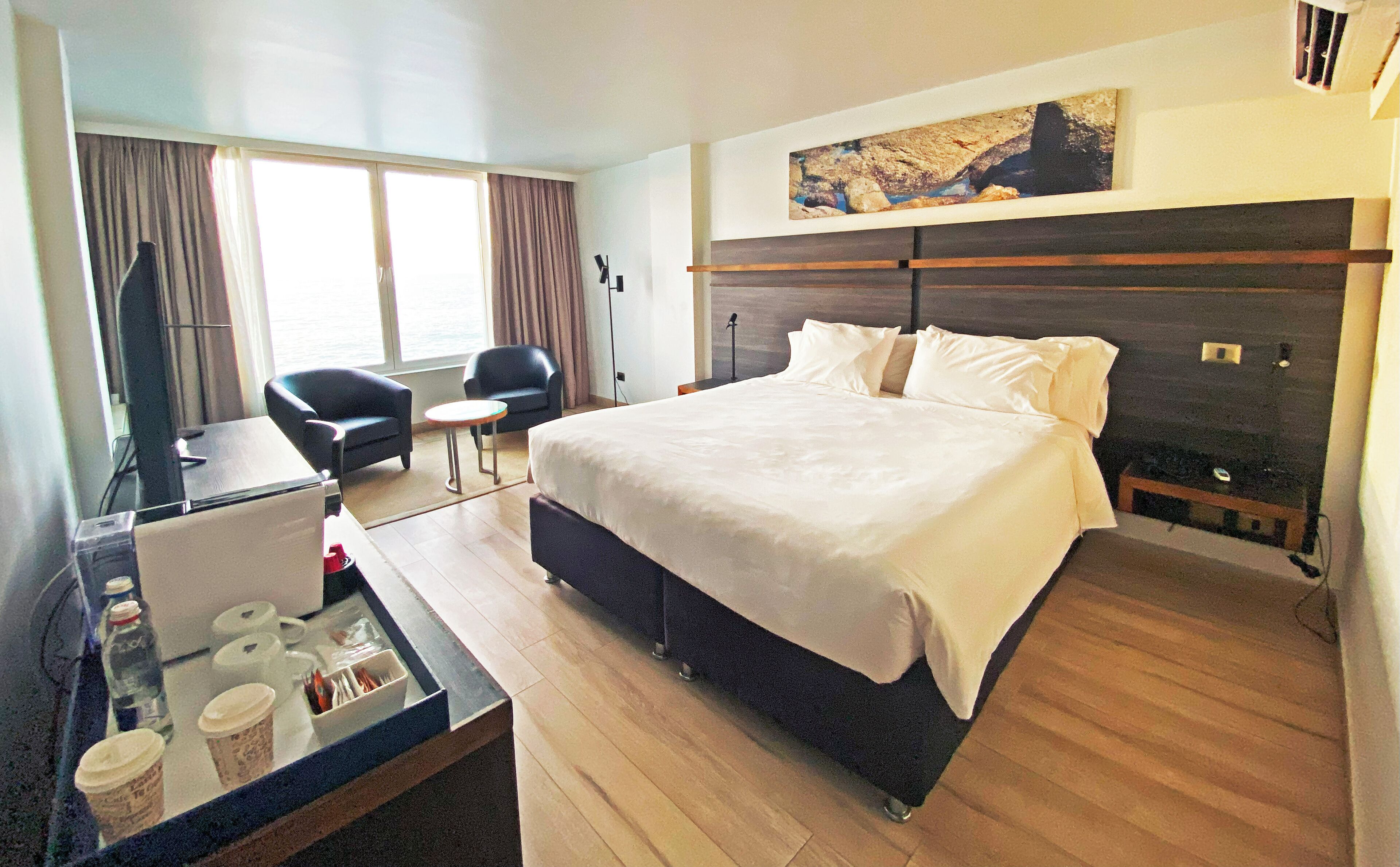 Een hotelkamer met een bed, twee stoelen, een bureau met een computer en koffiekopjes, en een schilderij aan de muur.