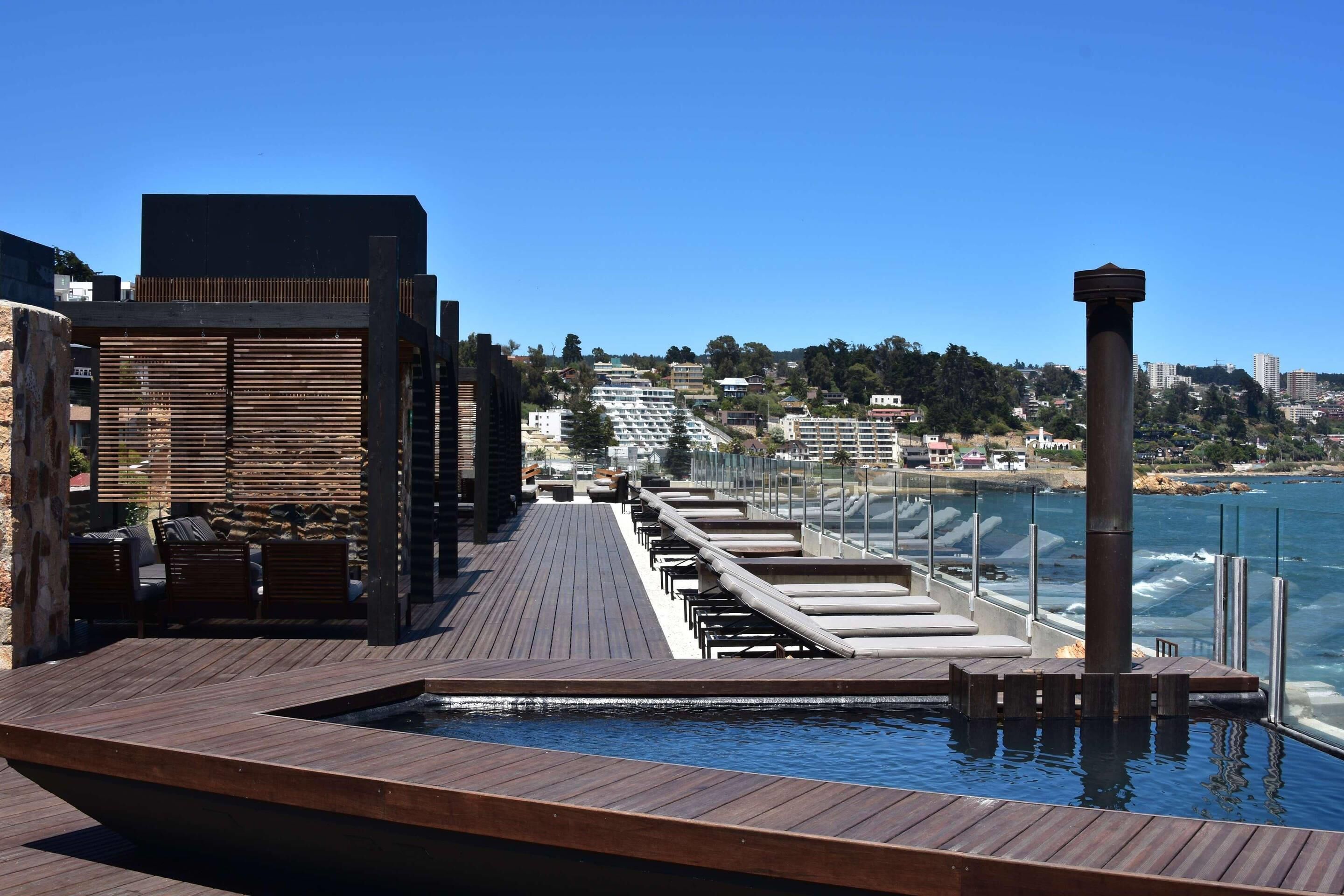Une terrasse en bois avec des chaises longues donnant sur une étendue d’eau, avec des bâtiments et un ciel dégagé en arrière-plan.