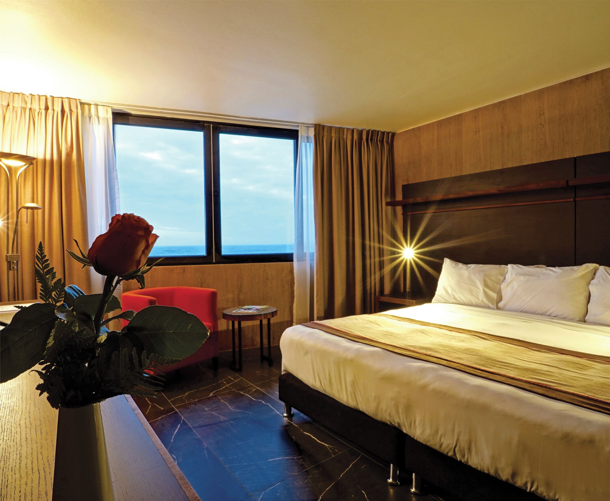 A hotel room with a large bed, a red chair, a small table, a vase with a rose, and a view of the sea through the window.