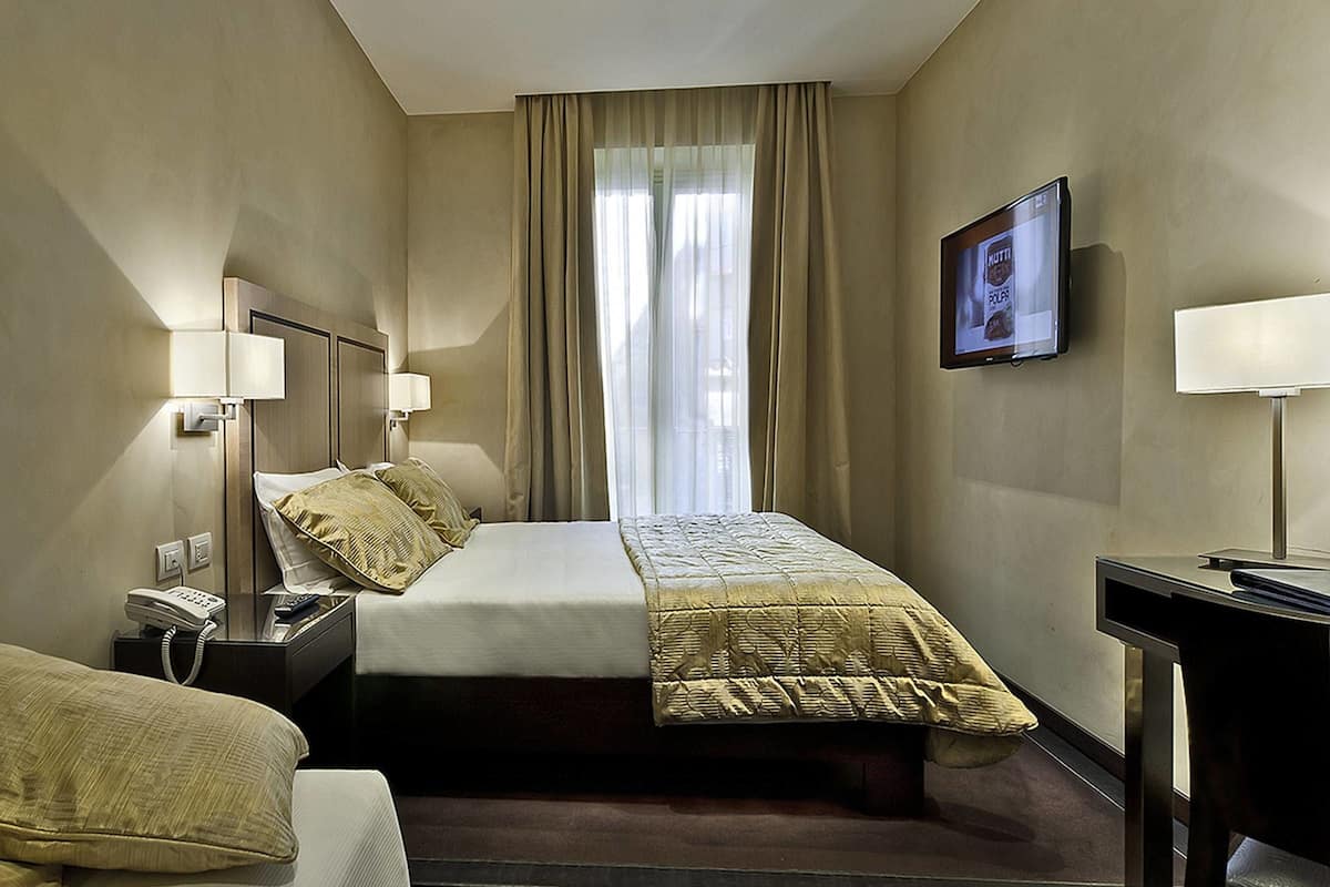 Habitación de hotel con cama, mesitas de noche, escritorio, silla, televisor y ventana con cortinas.