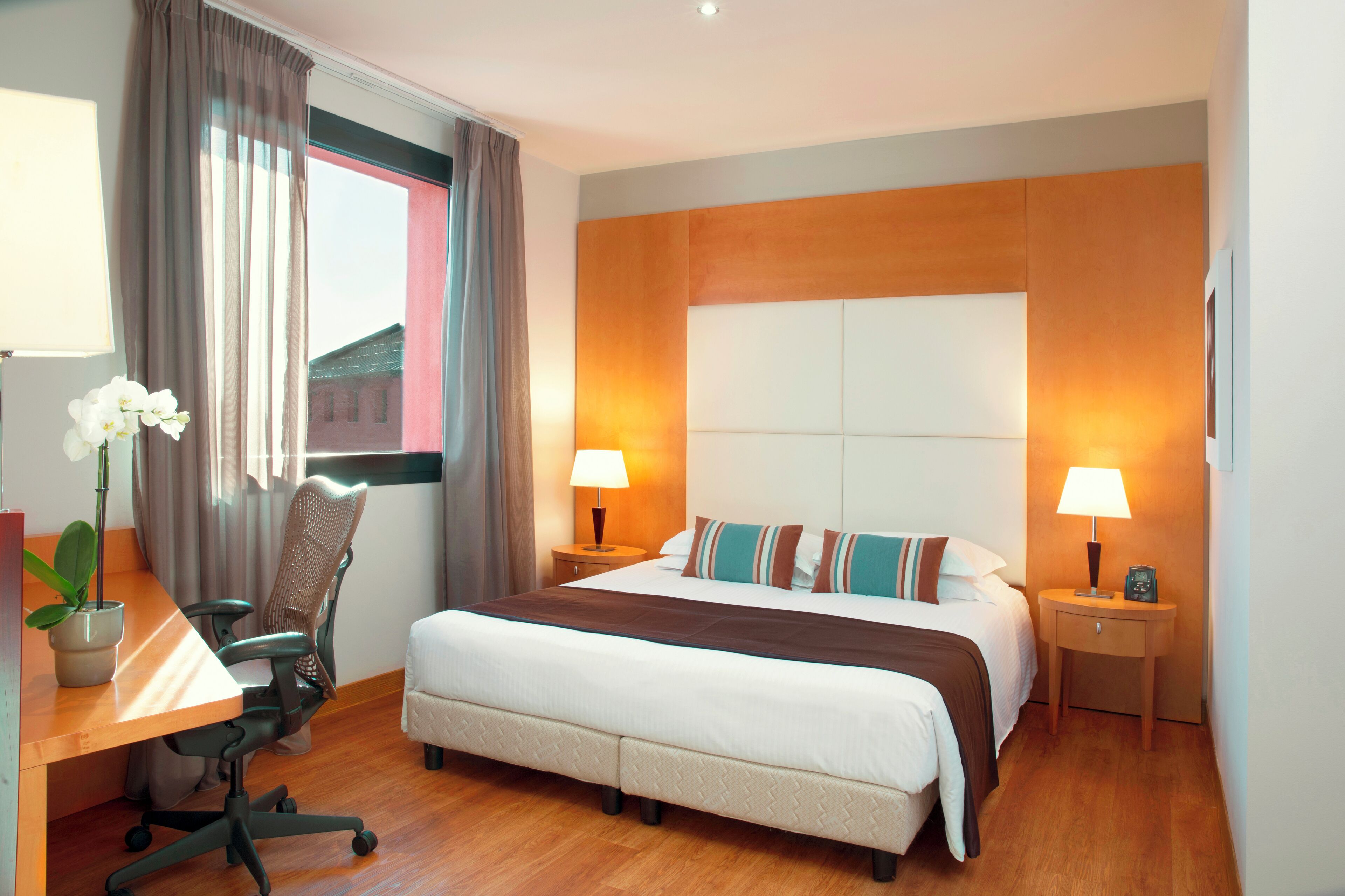 Habitación de hotel con una cama grande, dos mesitas de noche con lámparas, un escritorio con silla y una ventana con cortinas.