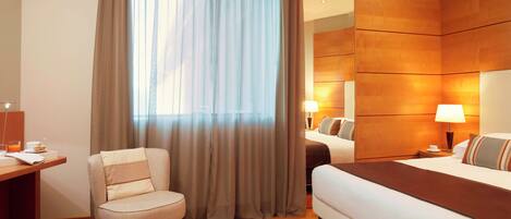 Una habitación de hotel con cama, una silla, escritorio y un amplio ventanal con cortinas.