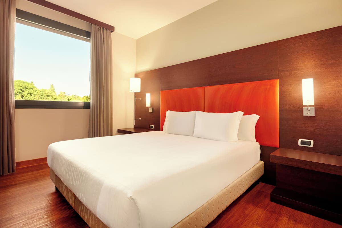 Una camera d'albergo con un letto grande, una testiera in legno, due comodini con lampade e una finestra con tende.