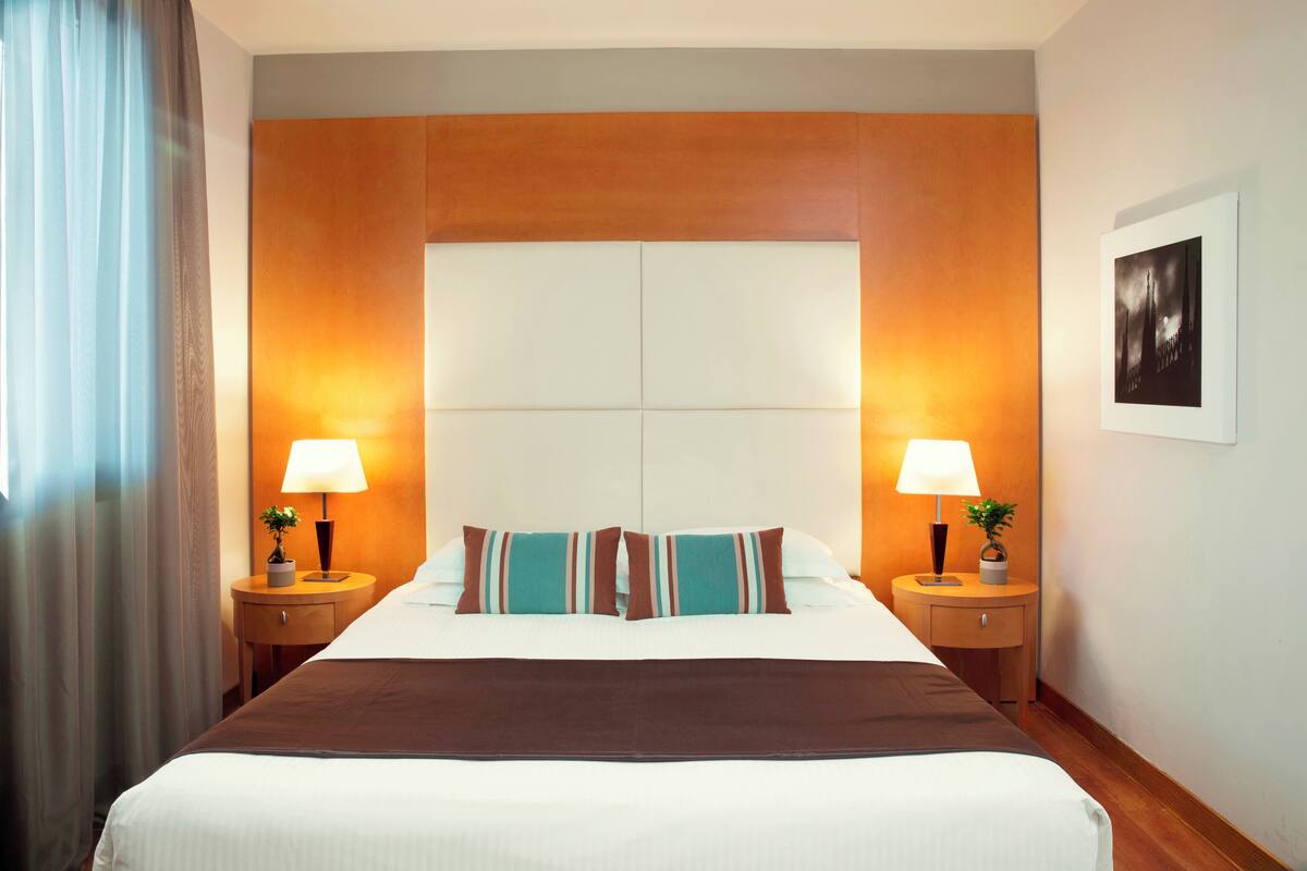Una camera d'albergo con un letto grande, due comodini con lampade, una testiera e un quadro appeso al muro.