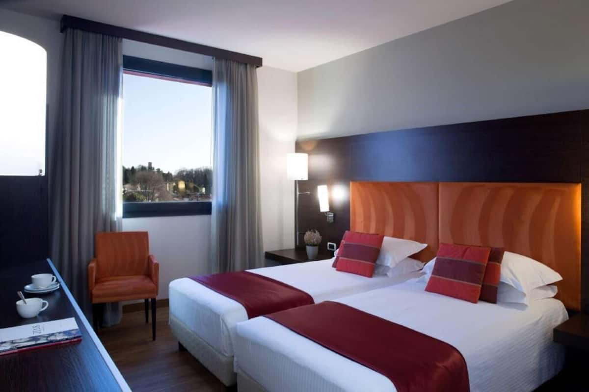 Una camera d'albergo con due letti, una scrivania con sedia, una finestra con tende e un comodino con lampada.
