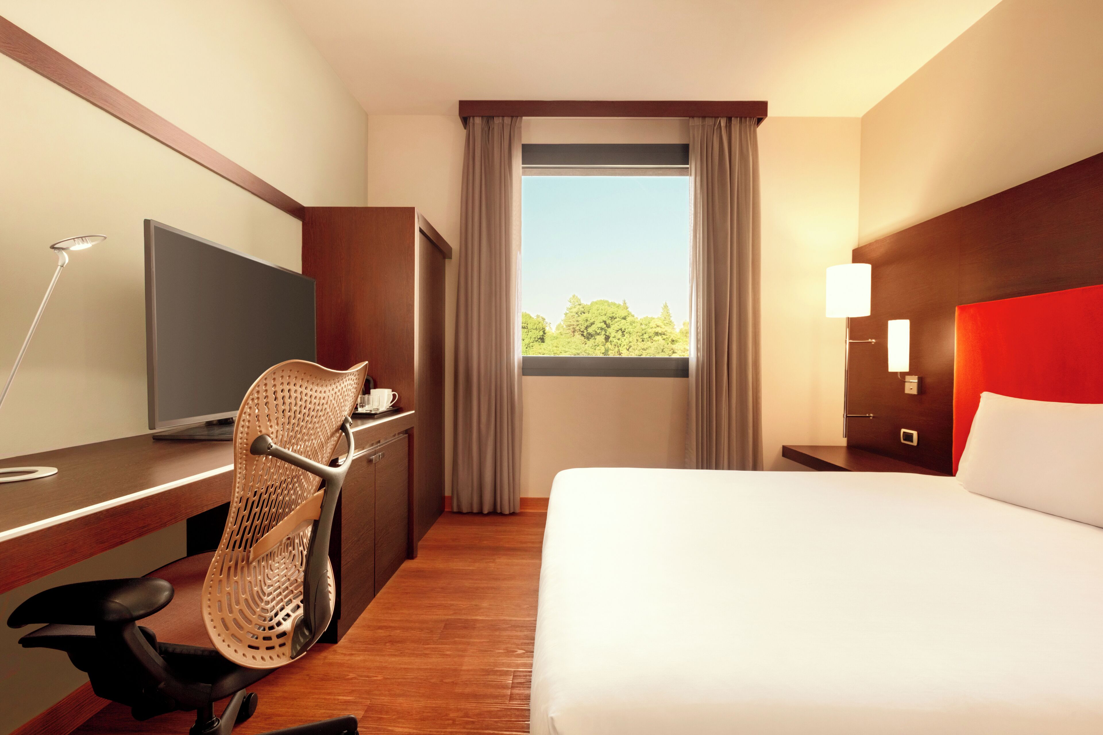 A hotel room with a bed, desk, chair, TV, and a window with curtains.