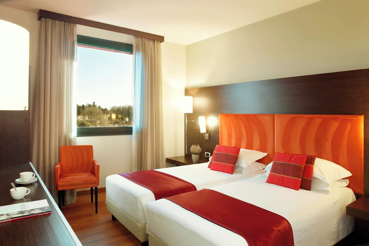 Una camera d'albergo con due letti, una scrivania con una sedia, una finestra con tende e un comodino con una lampada.