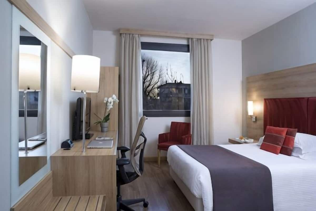 Una camera d'albergo con un letto, una scrivania con una lampada, una sedia, uno specchio e una finestra con tende.