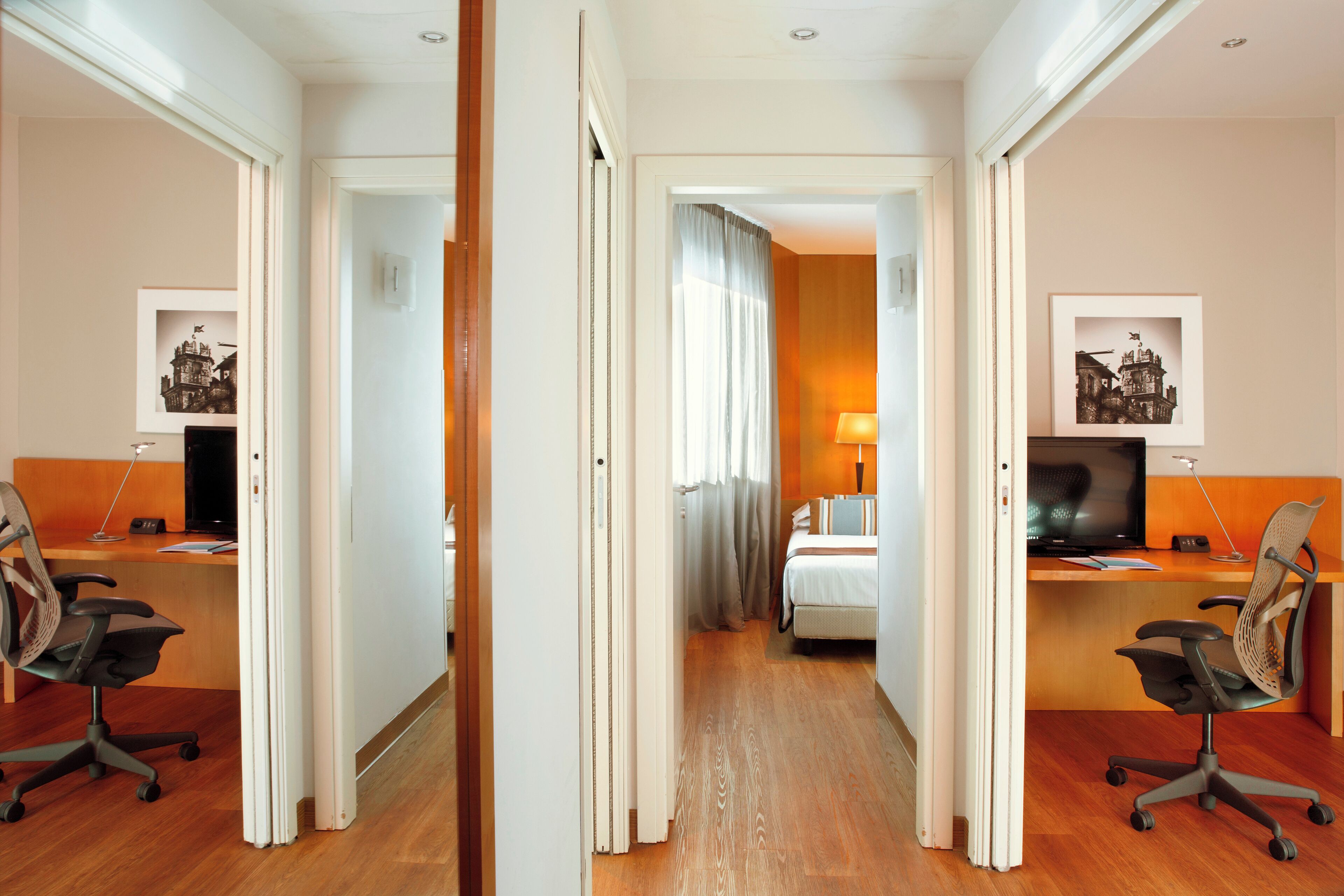 Een hotelkamer met een houten vloer, een bureau met een computer, een stoel, een bed en een raam met gordijnen.