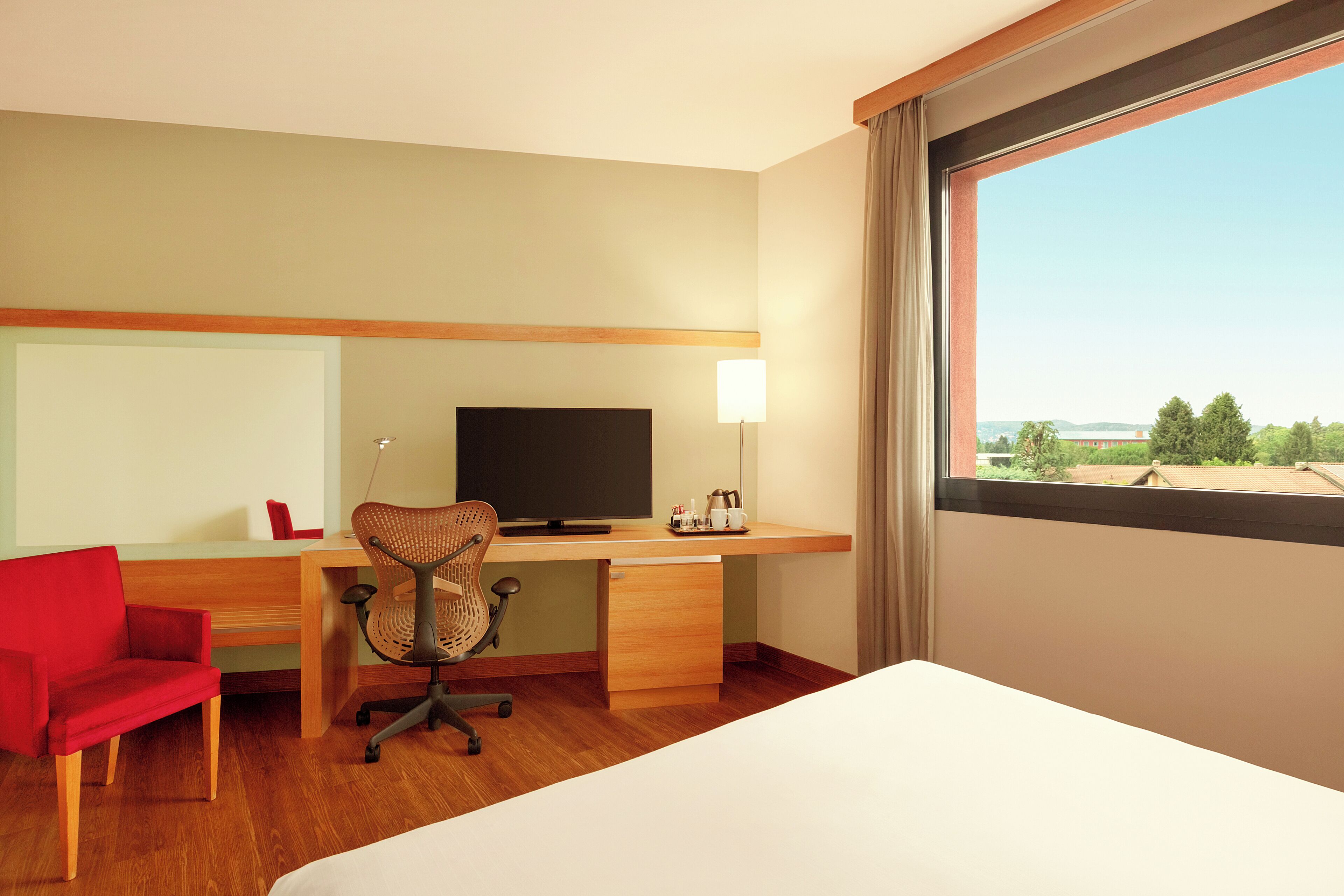 Una habitación de hotel con un escritorio de madera, televisor, una silla roja y una cama.