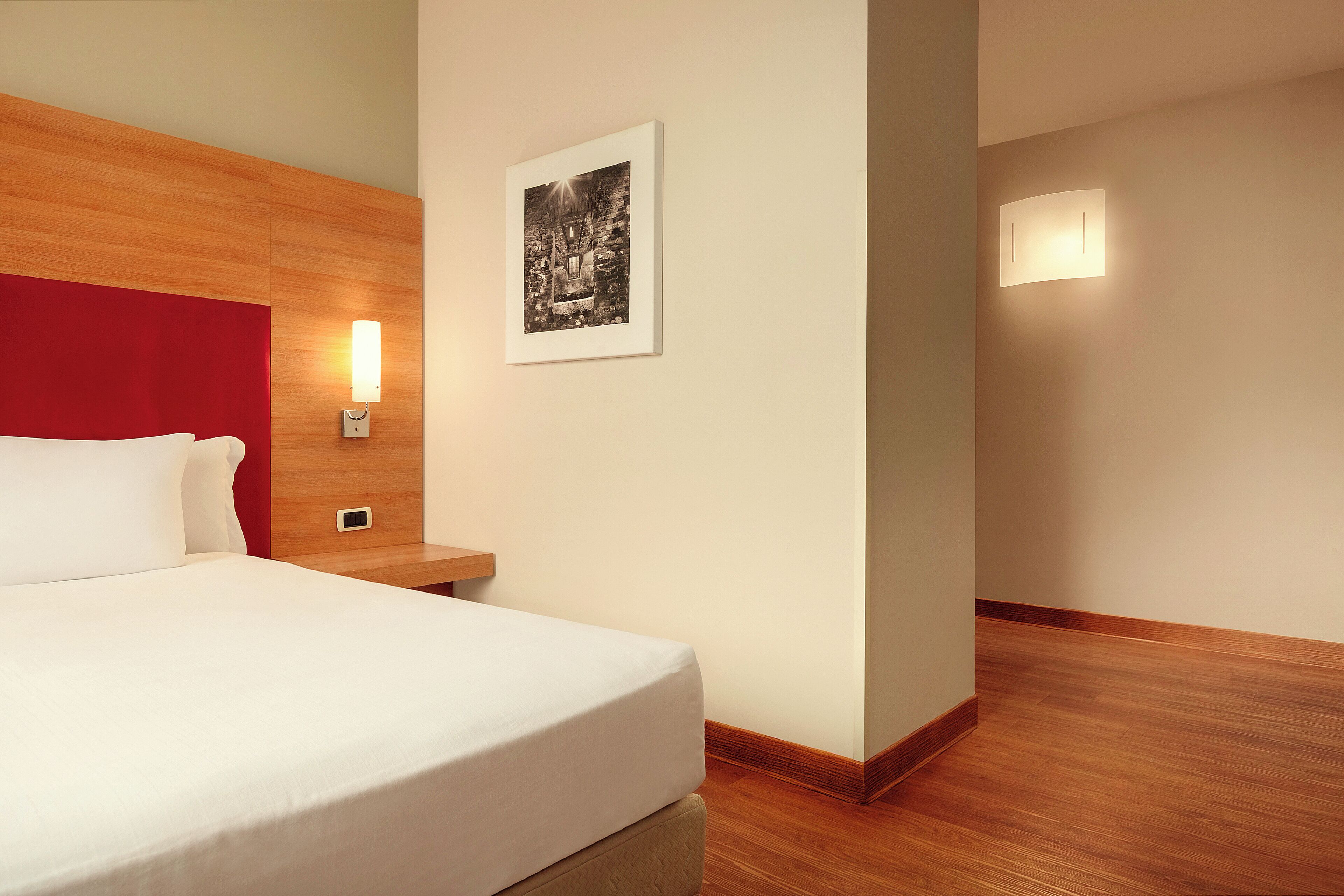 Habitación de hotel con cabecera de madera, cama con ropa de cama blanca, mesita de noche con lámpara y un cuadro en la pared.