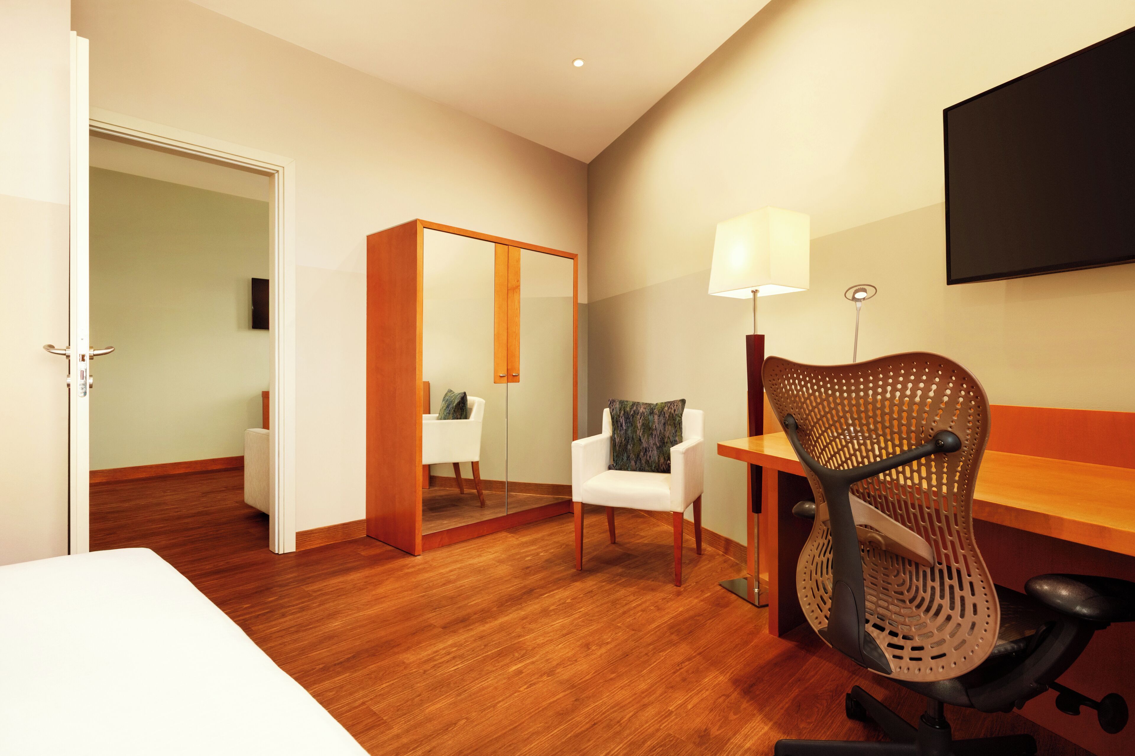 Ein Hotelzimmer mit Holzboden, einem Bett, einem Schreibtisch mit Stuhl, einem Spiegel und einem an der Wand befestigten Fernseher.
