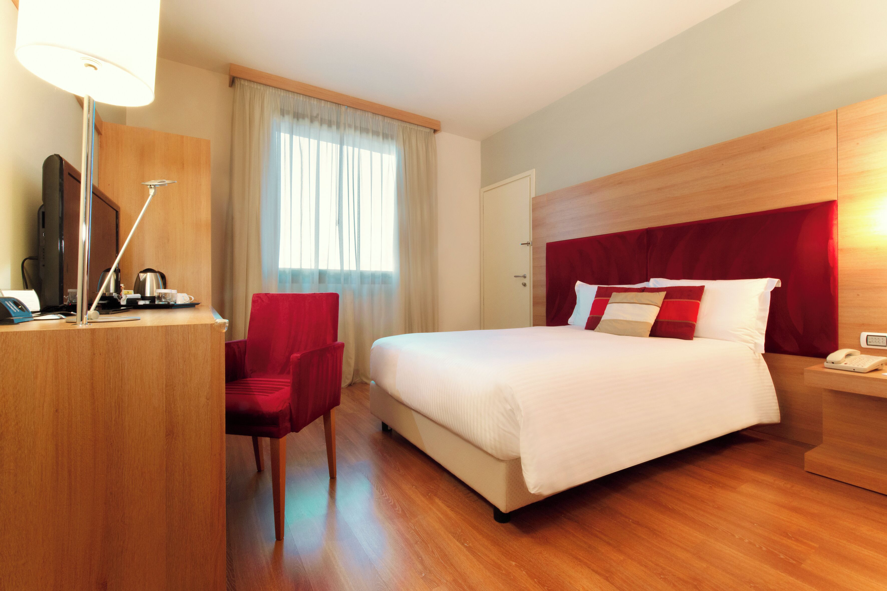 A hotel room with a bed, a desk with a telephone, a chair, a television, and a window with curtains.