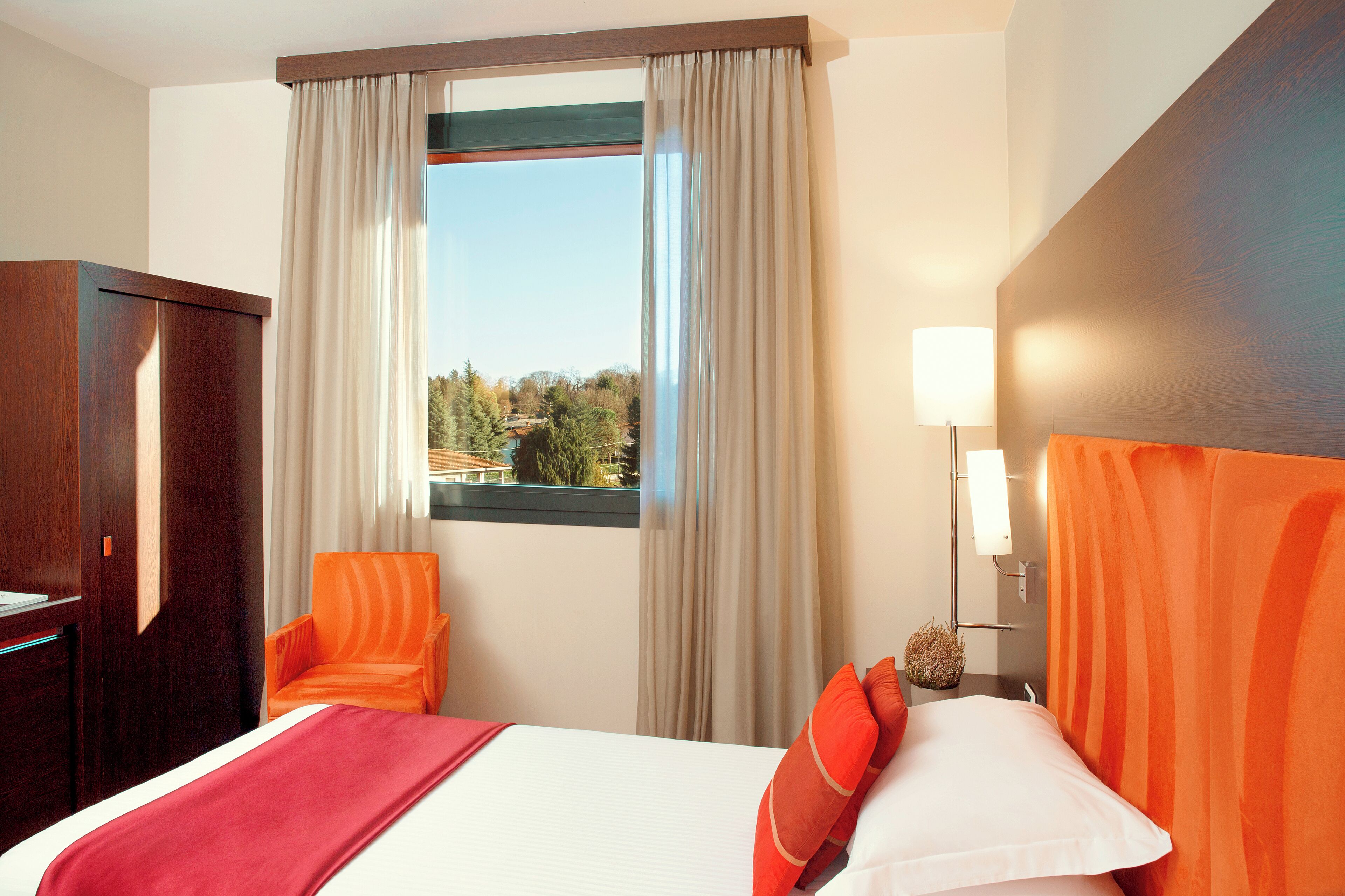A hotel room with a bed, an orange chair, a desk, a lamp, and a view of the outdoors.