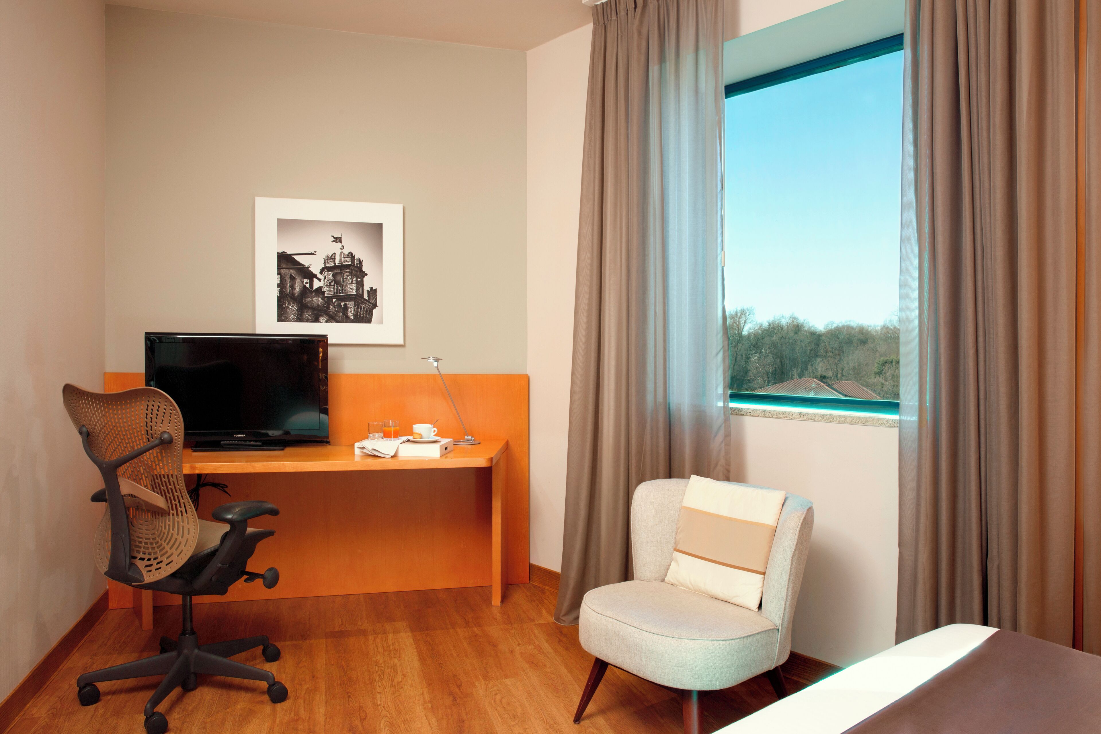 Una camera d'albergo con una scrivania in legno, una televisione, una sedia, un letto e una finestra con tende.