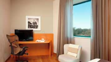 Una camera d'albergo con una scrivania in legno, una televisione, una sedia, un letto e una finestra con tende.