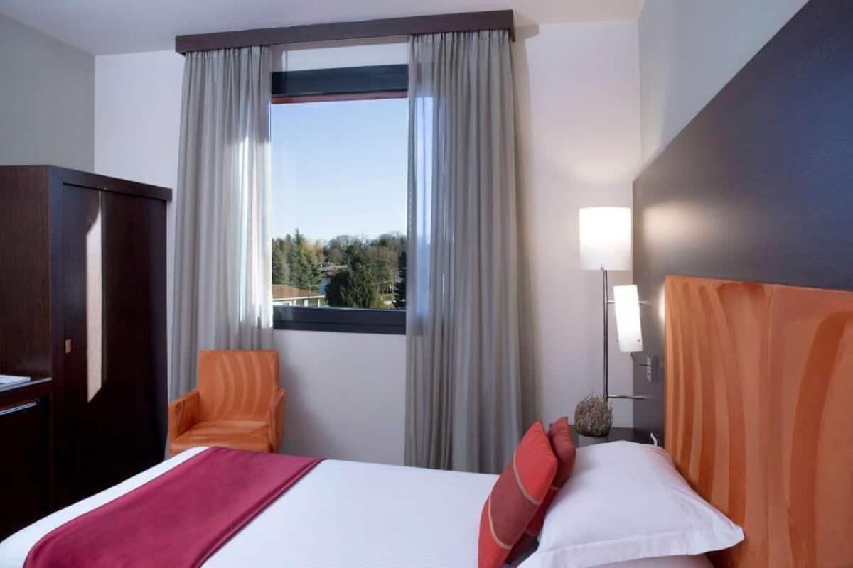 Una camera d'albergo con un letto, una sedia, una scrivania, una lampada e una finestra con tende.