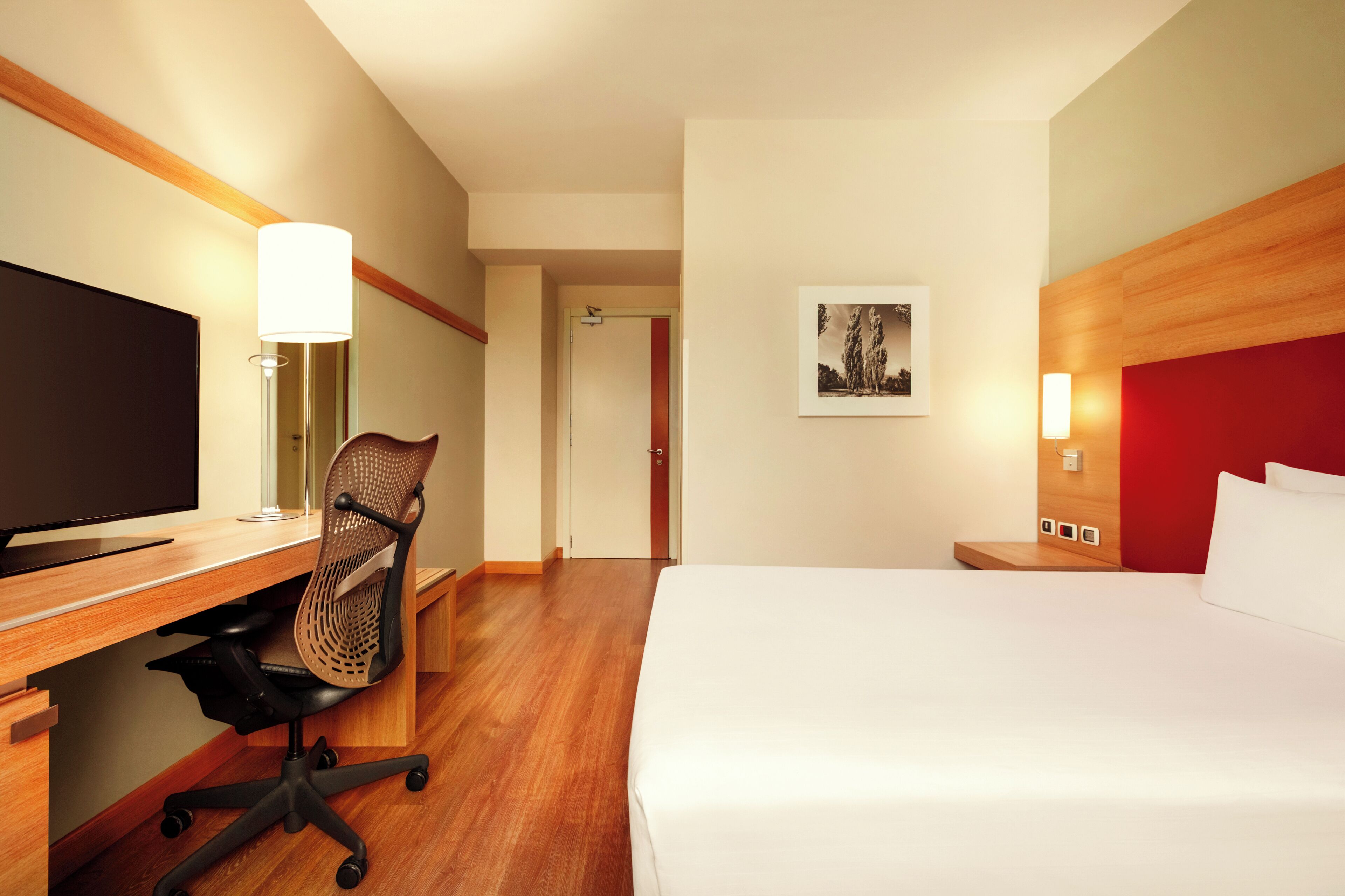 A hotel room with a bed, desk, chair, television, and artwork on the wall.