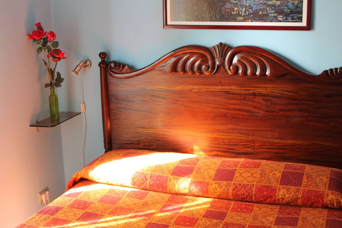 A wooden bed with a patterned bedspread and a vase with red flowers on a bedside table.