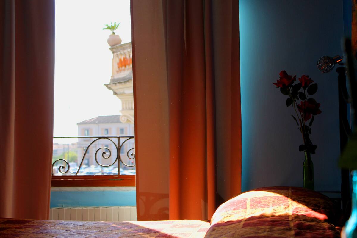 A room with a bed, a window with curtains, a vase with flowers, and a view of buildings outside.