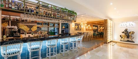 Zona de bar con estantes llenos de botellas de licores, copas de vino colgando y taburetes de mimbre blanco.